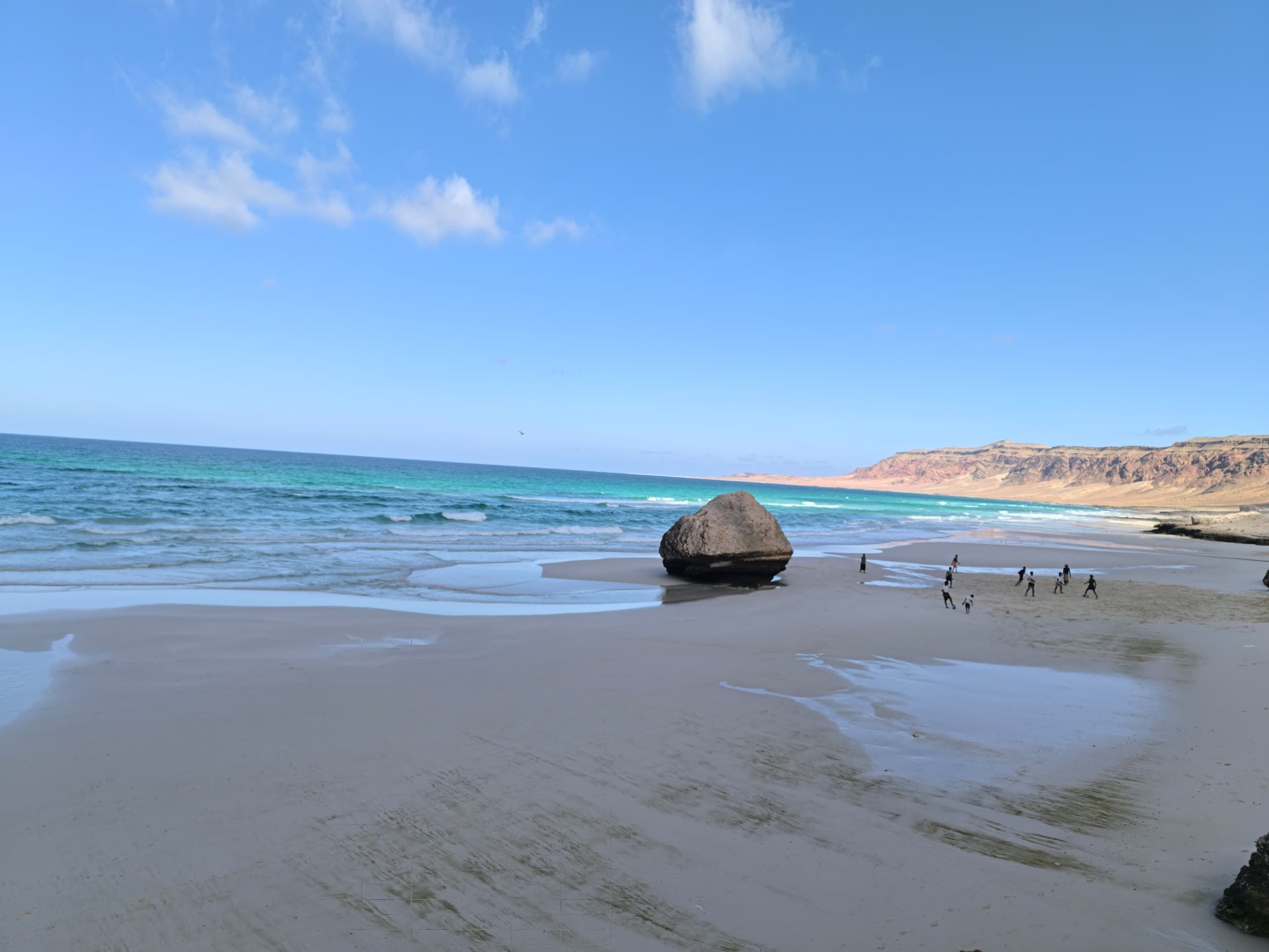 A single large boulder on a wet tidal flat, with small figures walking beyond it