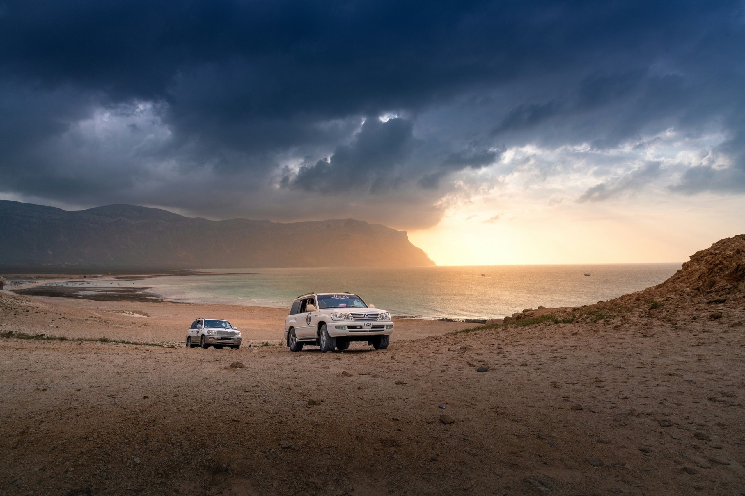Two white four-wheel-drive vehicles on a gravel rise above a beach under heavy storm cloud