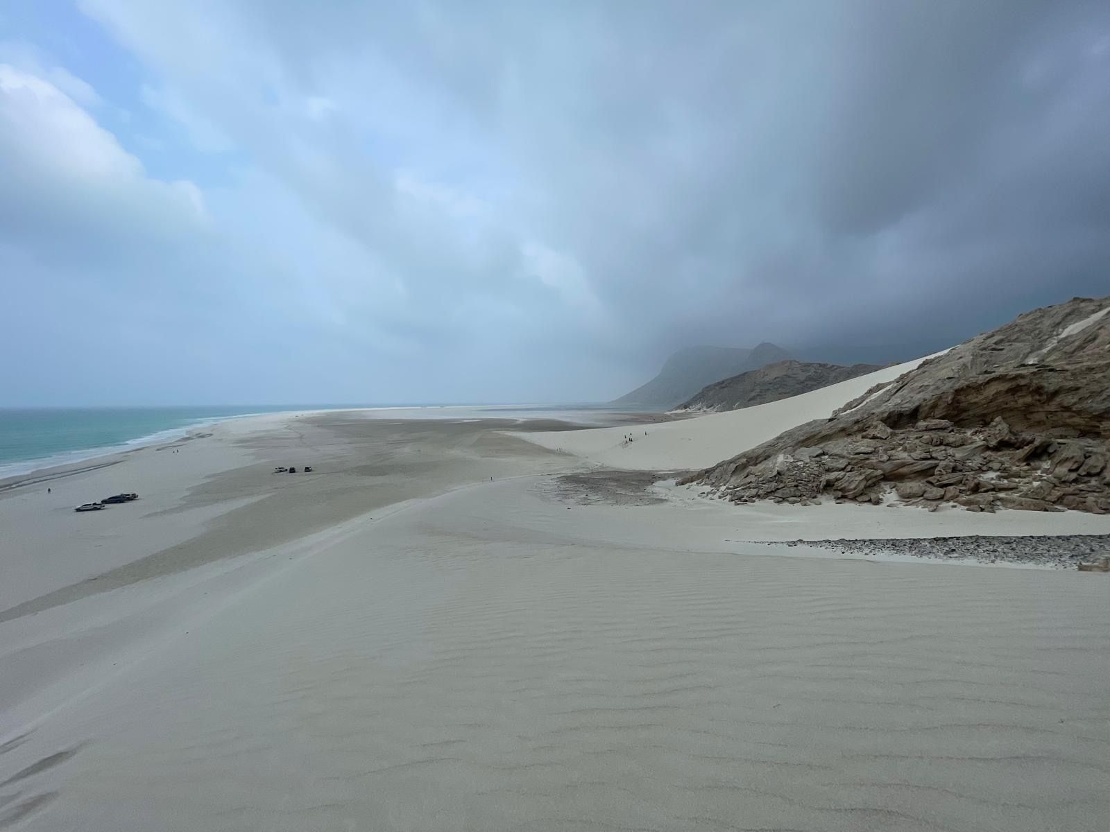 A large dune meeting rock under dark cloud, vehicles small in the distance