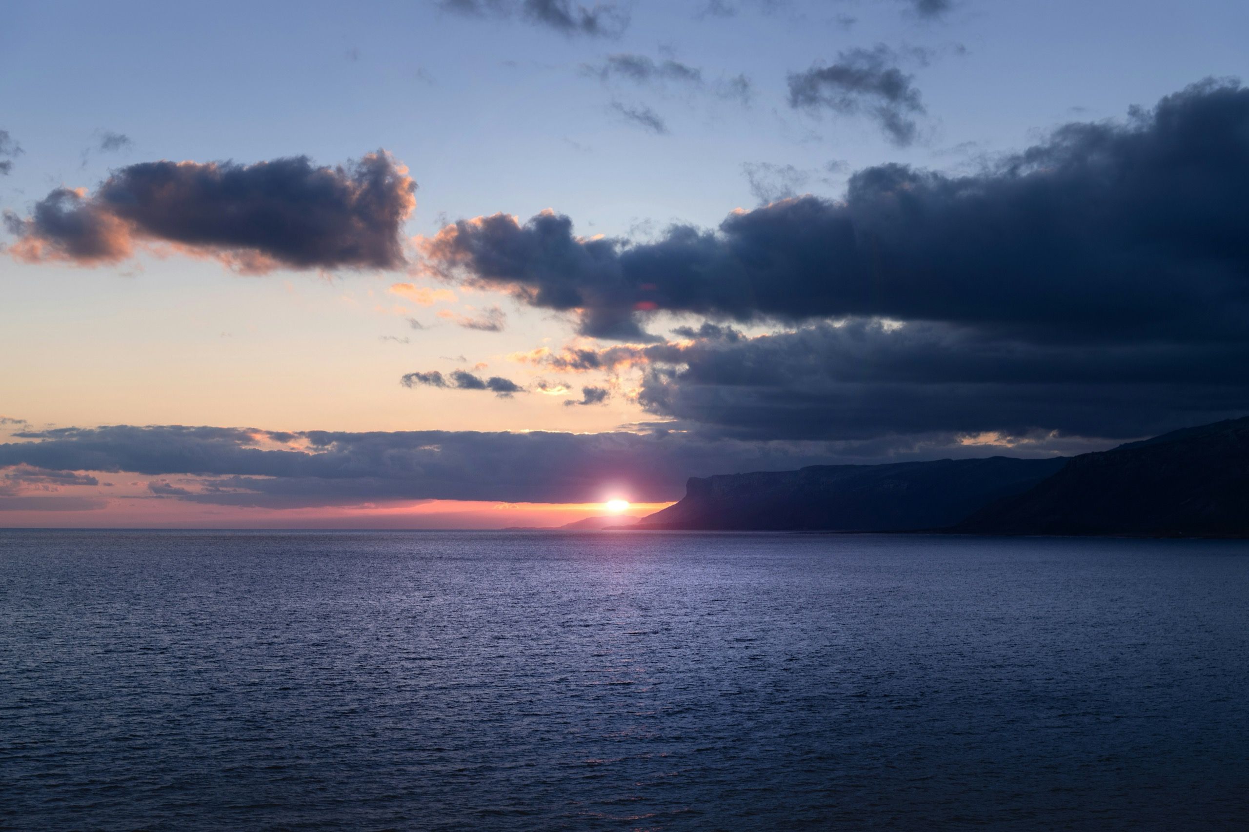 The sun low on the horizon over open sea, with a headland silhouetted to one side