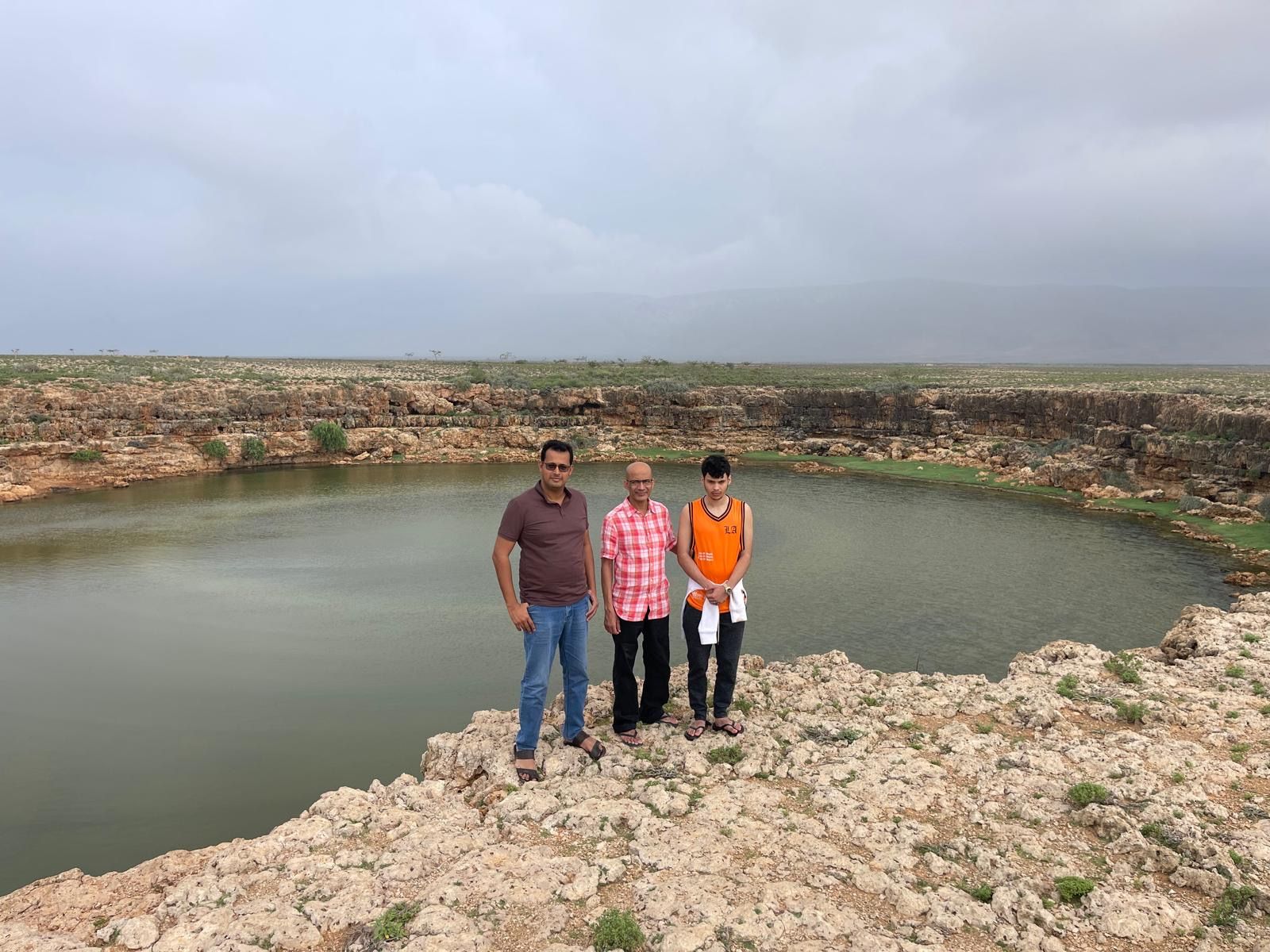 Three men standing at the edge of a natural rock pool under a grey sky