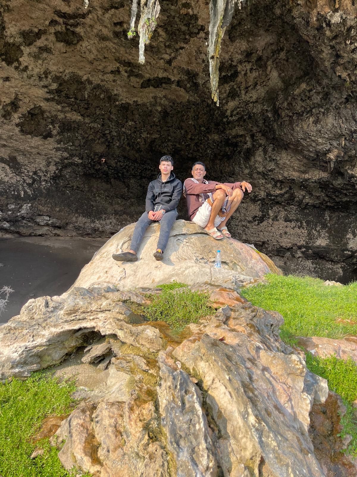 Two men sitting on a rock at the mouth of a cave, dark overhang above them