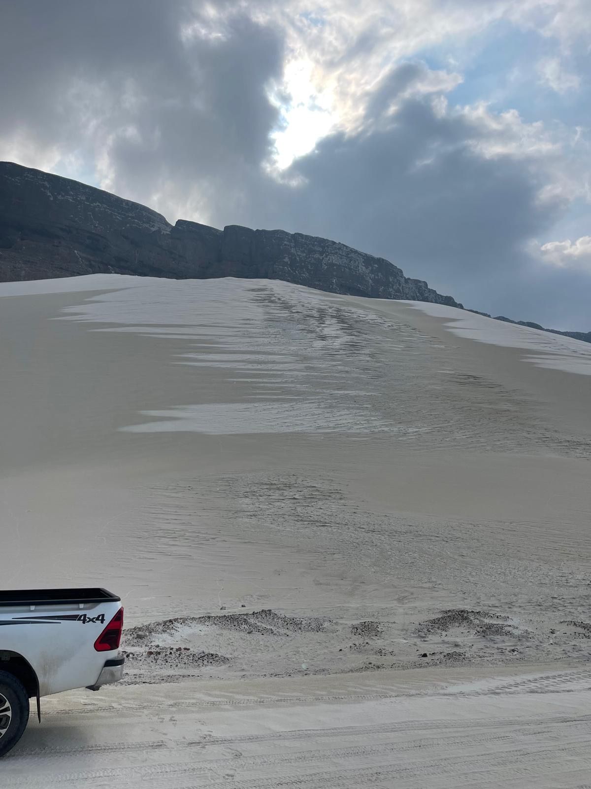 A white dune climbing to meet a dark cliff, a four-wheel-drive parked below